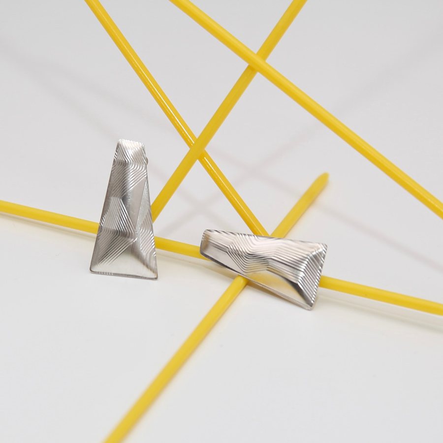 Pair of silver trapezoid stud earrings with fine guilloché lines, resting on yellow rods
